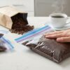 キッチンのカウンターで、コーヒー粉が入ったジップロックを上から手で押さえて空気を抜いている様子。背景には開封されたコーヒー豆の袋と、湯気が立つコーヒーカップが置かれている。